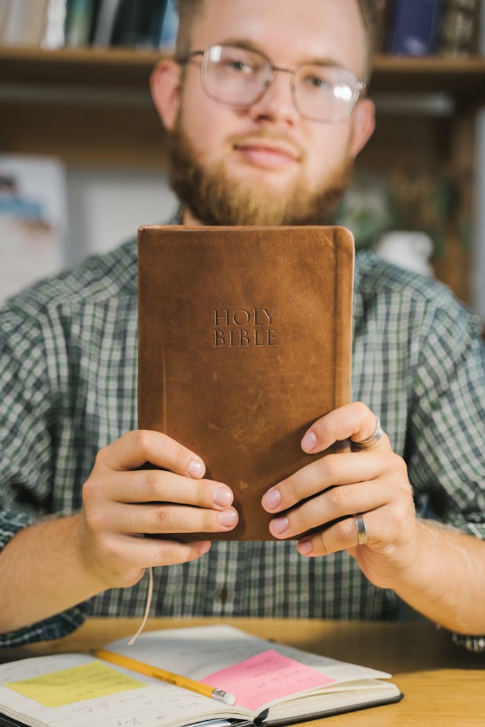 Bearded man with glasses holding a Holy Bible in a study environment.