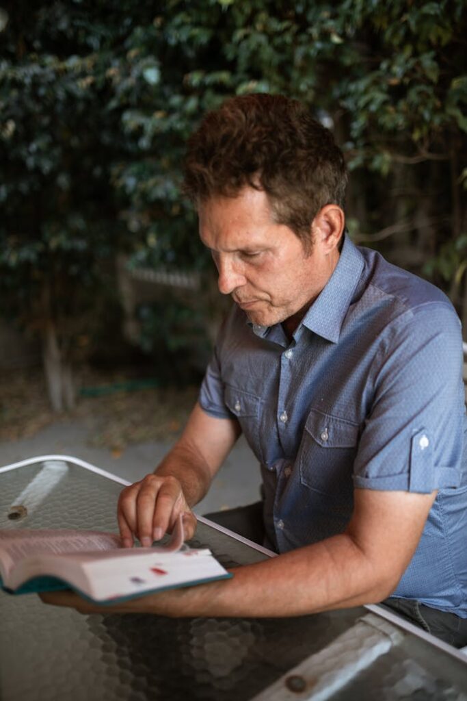 Adult man reading a book outdoors at a table surrounded by greenery.